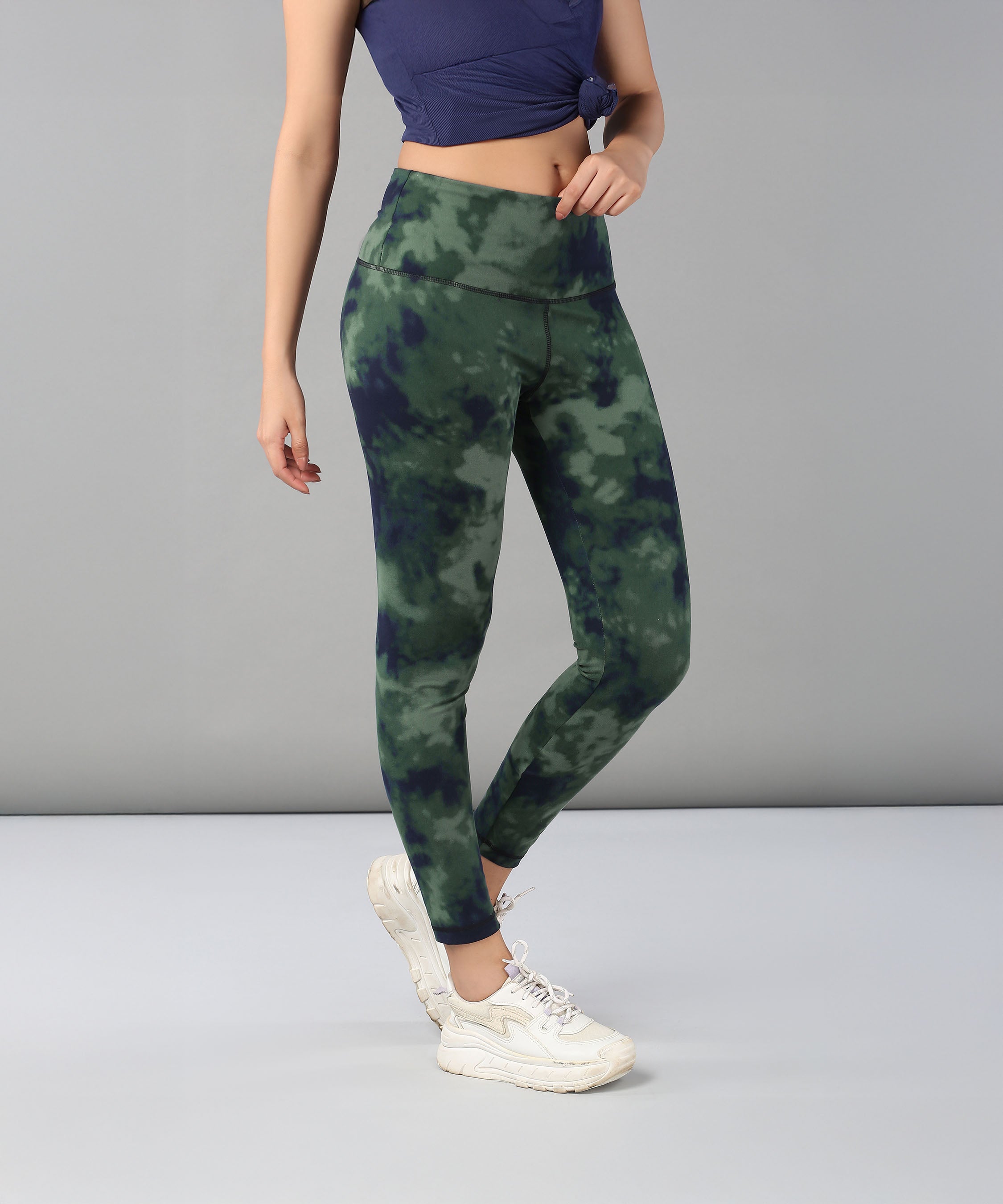 A woman models Tie & Dye Leggings in green stretchy fabric, paired with white sneakers and a blue top, standing against a plain gray background—ideal for enhancing your workout wardrobe.