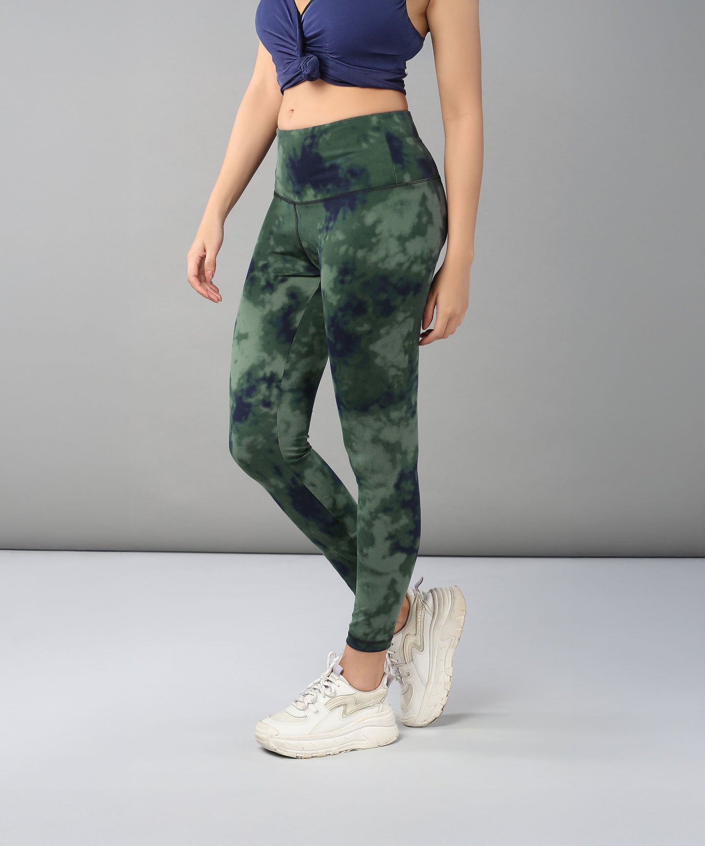 A woman models the Tie & Dye Leggings in green, made from stretchy fabric, paired with a navy crop top and white sneakers against a gray background—ideal for updating your workout wardrobe.