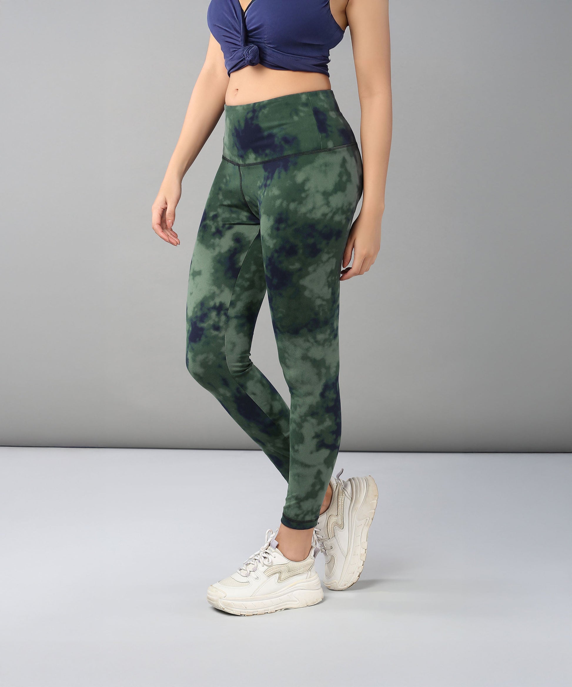 A woman models the Tie & Dye Leggings in green, made from stretchy fabric, paired with a navy crop top and white sneakers against a gray background—ideal for updating your workout wardrobe.