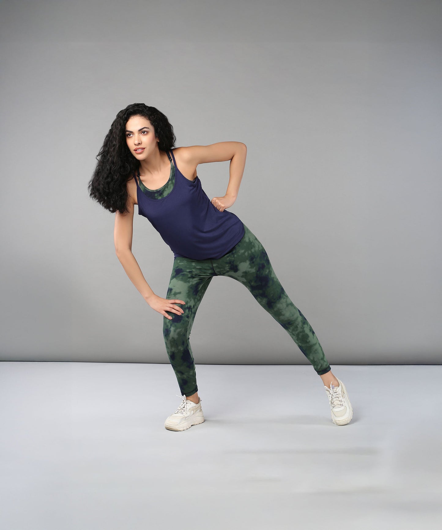 A woman in athletic wear stretches sideways against a plain grey background, highlighting Tie & Dye Leggings that bring vibrant color and flair to her workout wardrobe.