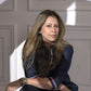 A woman with long hair sits indoors wearing the Accented Set, a dark batwing sleeve shirt and watch, as light and shadow dance on the wall behind her.