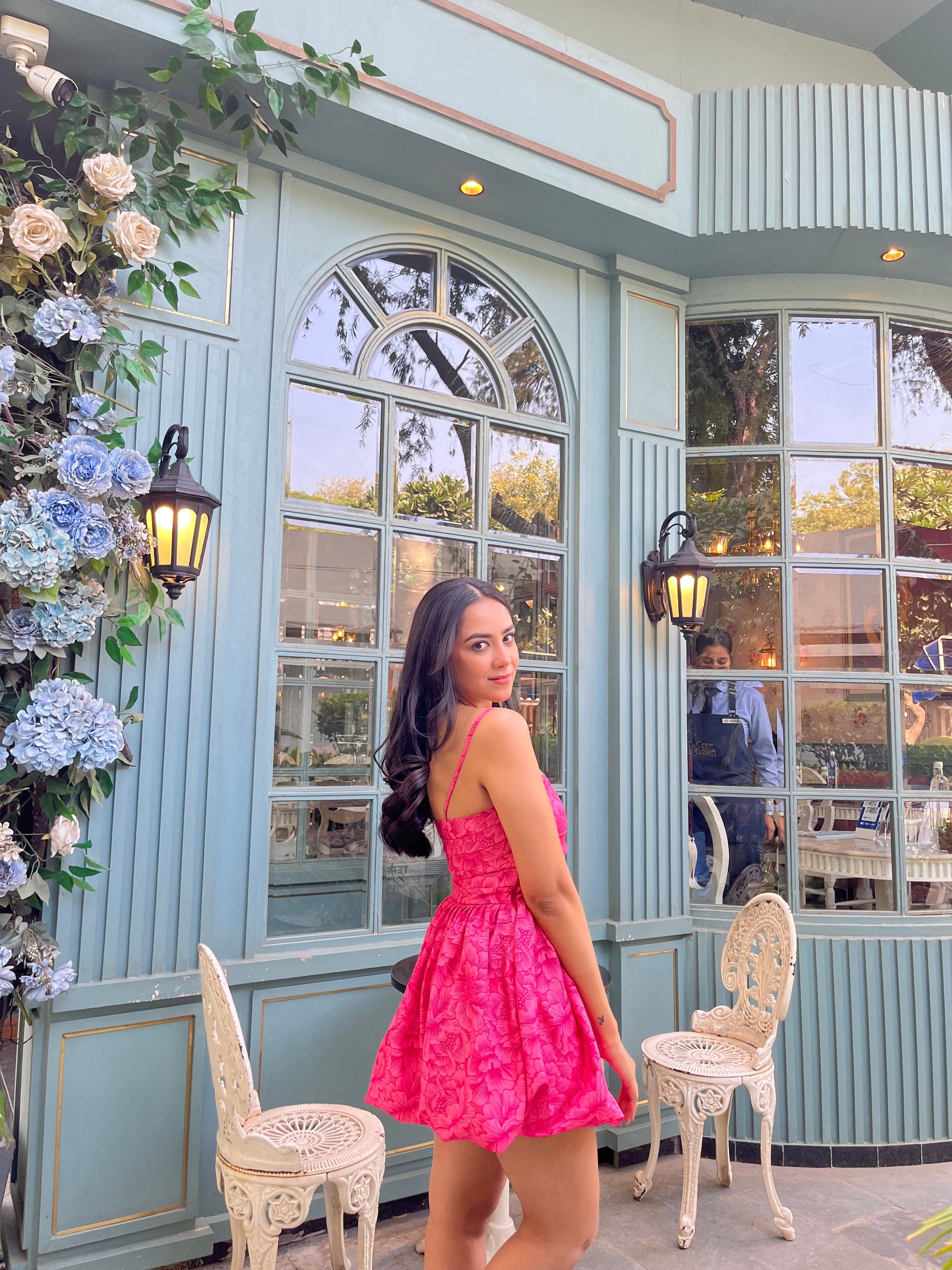 Woman in pink Barbie balloon dress posing by teal cafe with floral decor vintage chairs and arched windows