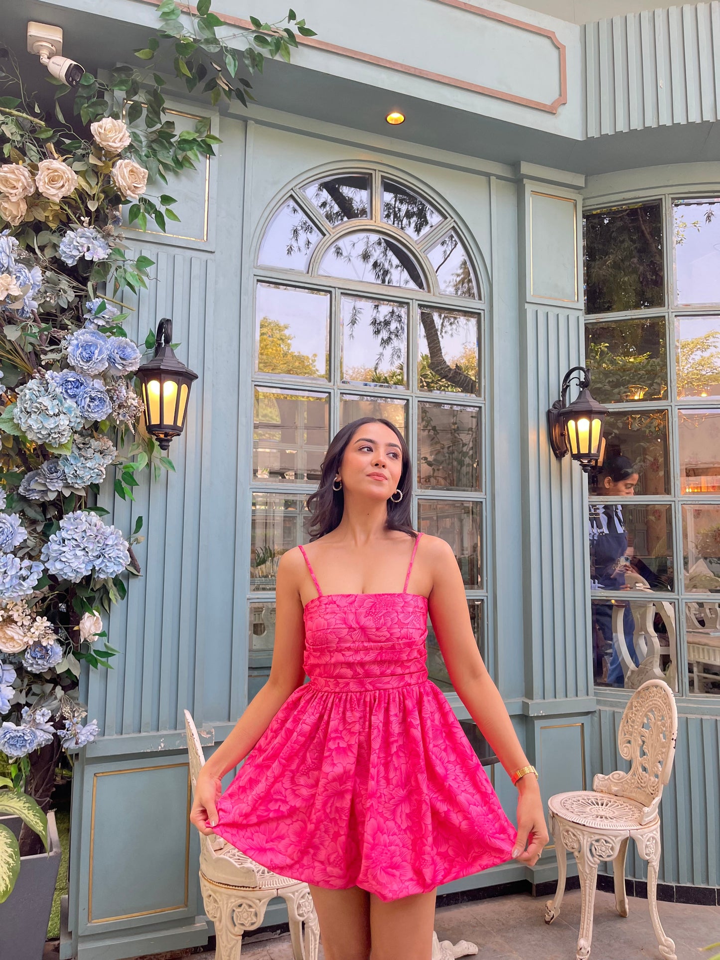 Woman modeling Stylera Barbie balloon dress in bright pink short fit and flare with thin straps by floral cafe backdrop