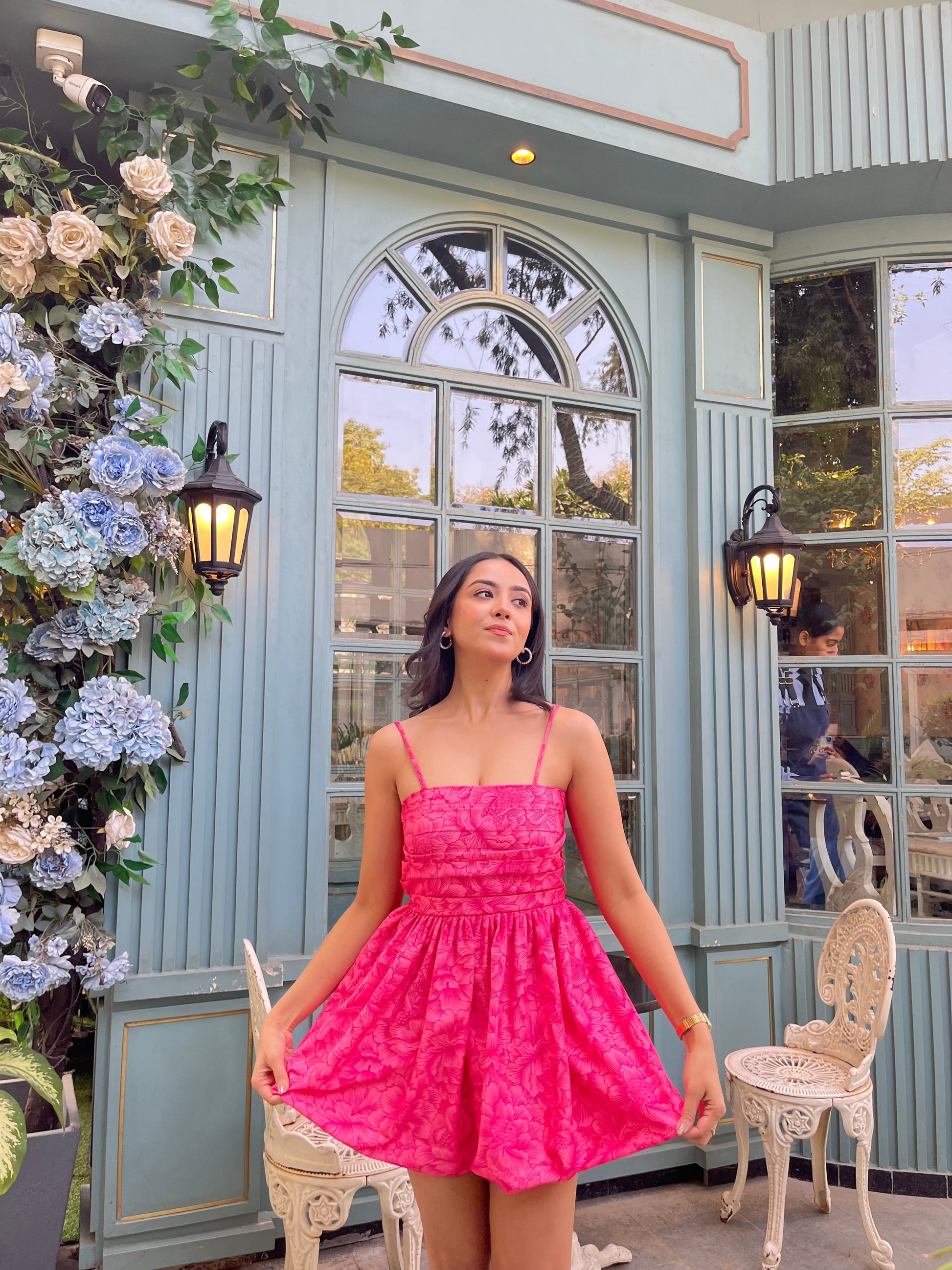 Woman modeling Stylera Barbie balloon dress in bright pink short fit and flare with thin straps by floral cafe backdrop