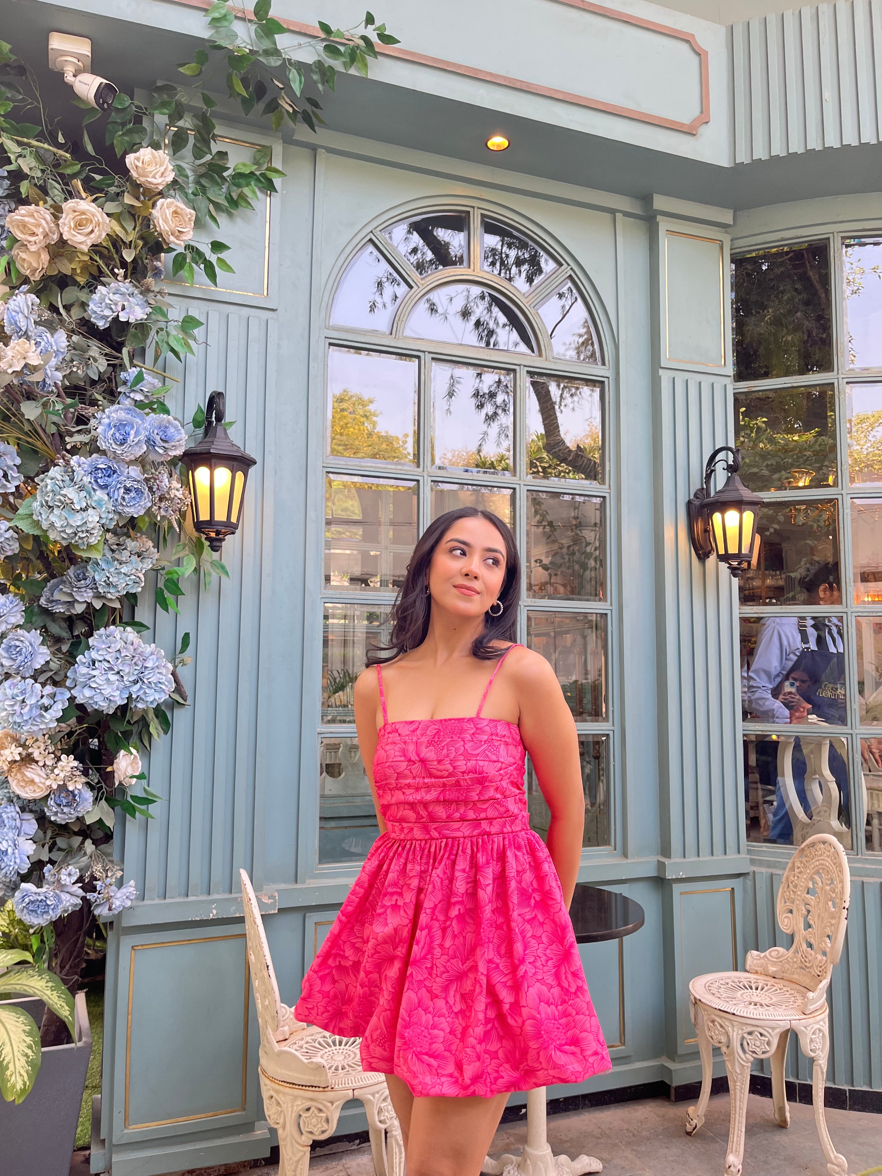 Model wearing a hot pink Barbie balloon dress with thin straps posed by pastel cafe window