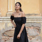 Model wearing a black off shoulder short dress with puff sleeves against a textured outdoor background