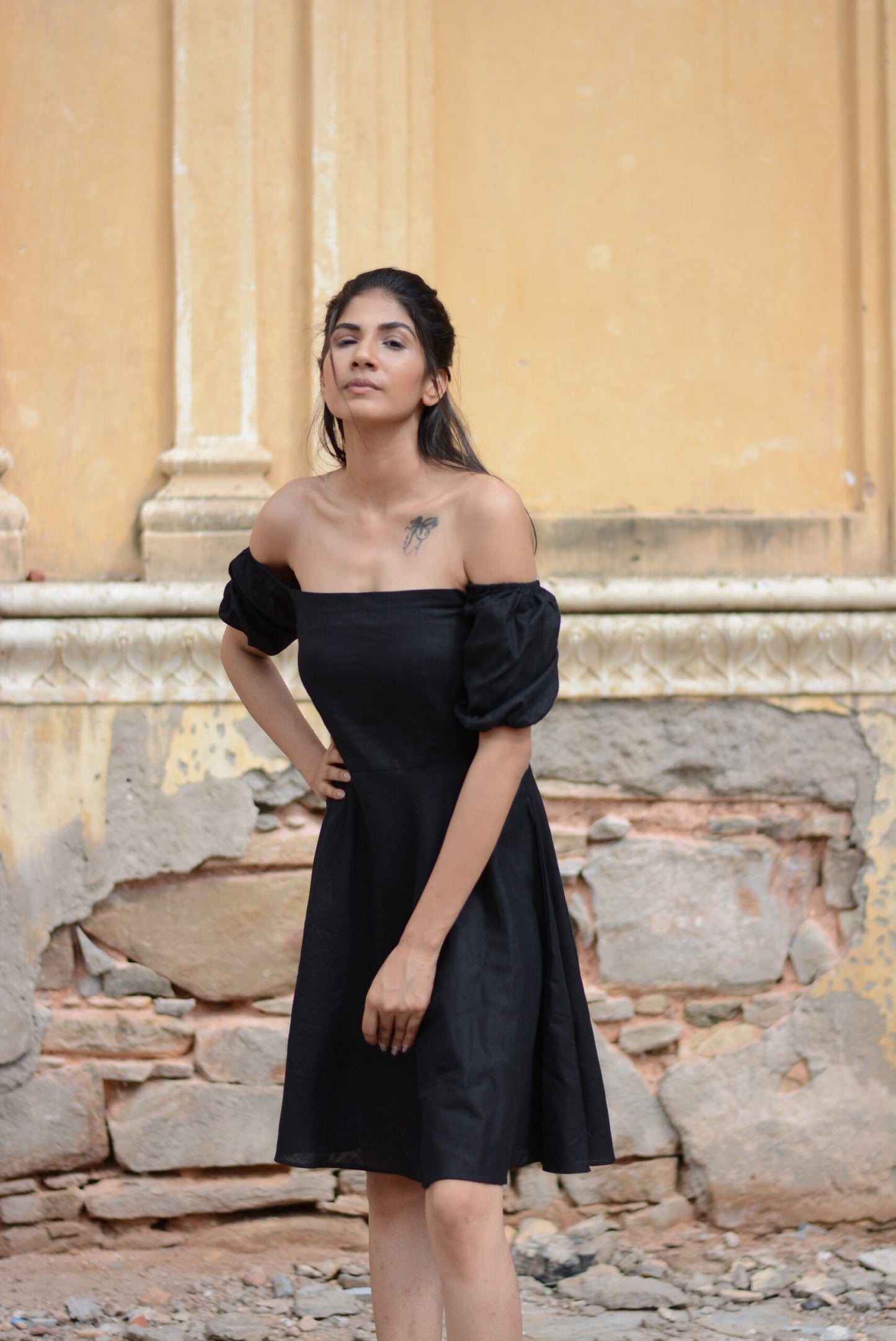 Model wearing a black off shoulder short dress with puff sleeves against a textured outdoor background