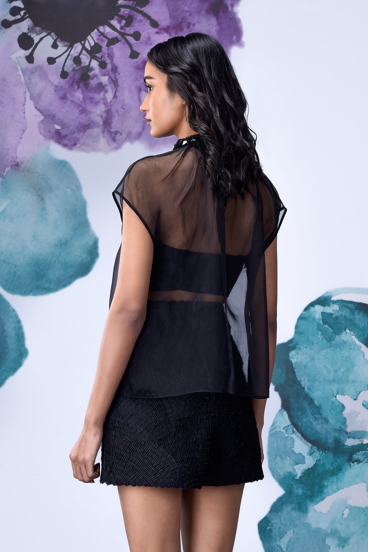 Back view of model wearing black organza top for women layered over bandeau with textured mini skirt