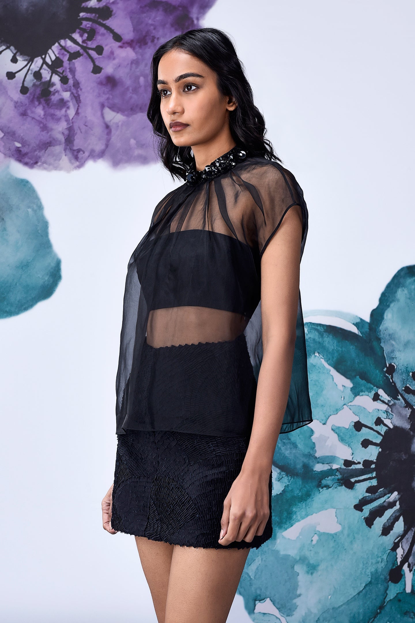 Model wearing a semi sheer black organza top for women with beaded collar over bandeau and lace mini skirt
