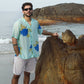 Man wearing a blue oversized shirt for men standing by the beach with rocks and ocean in the background