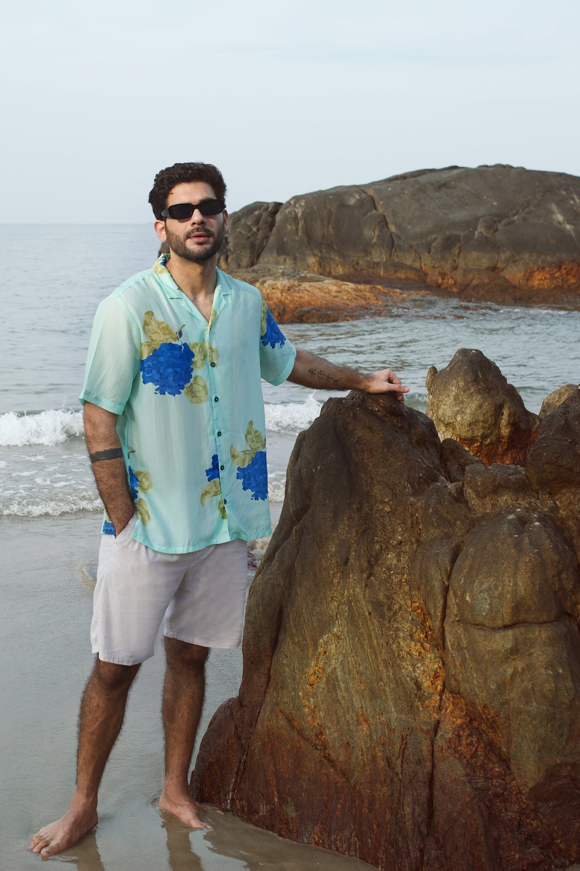 Man wearing a blue oversized shirt for men standing by the beach with rocks and ocean in the background