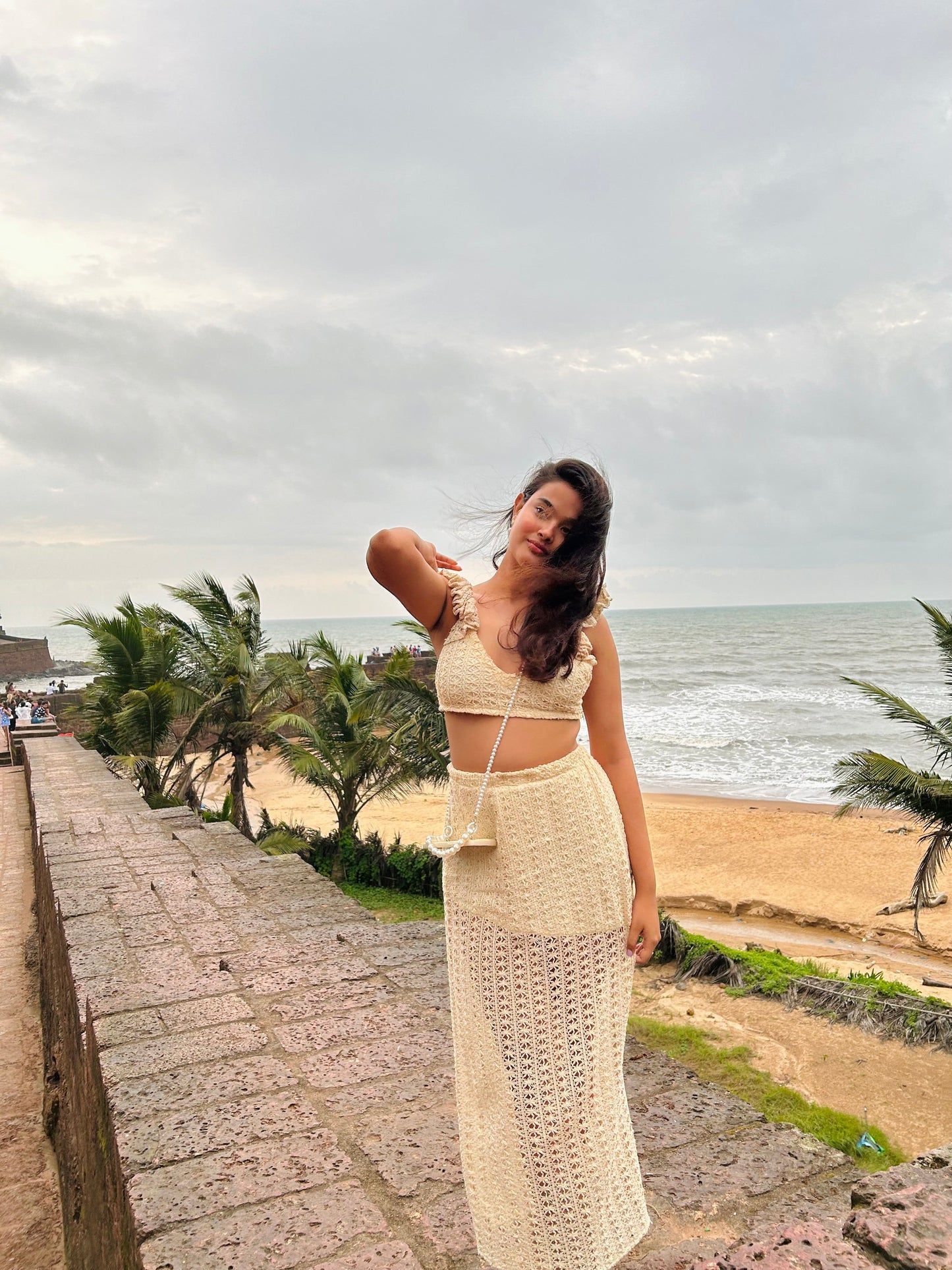Woman in cream crochet two piece set on sea wall with palm trees and ocean breeze trendy seaside vacation outfit