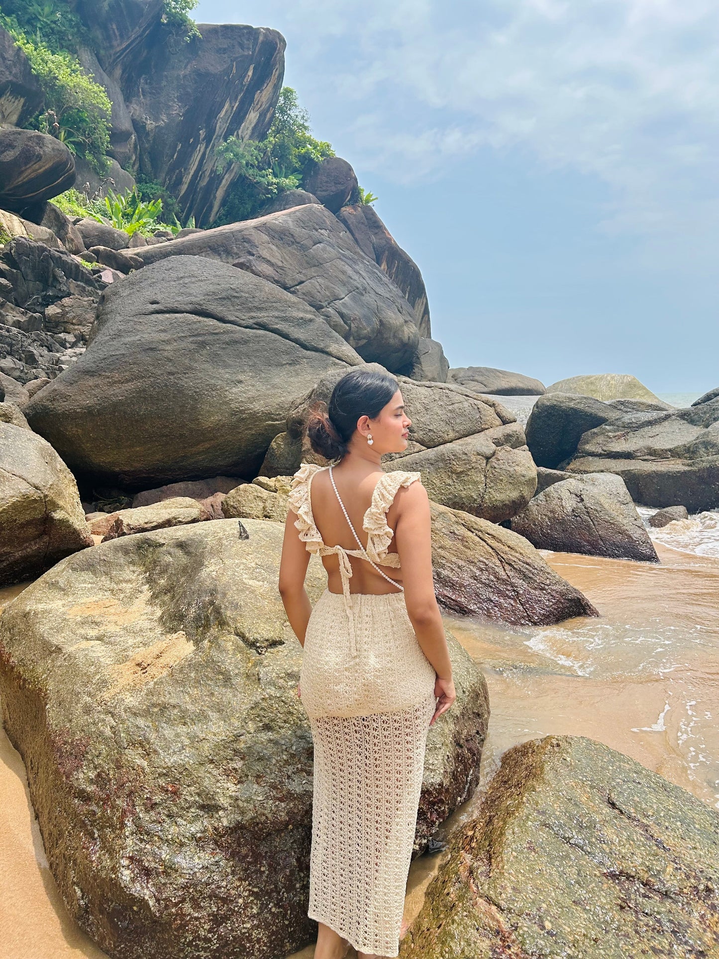 Woman wearing cream crochet open back dress on rocky beach showcasing Stylera trendy seaside vacation outfit