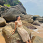 Stylera model in beige crochet two piece set sitting on coastal rocks and sand in a trendy seaside vacation outfit