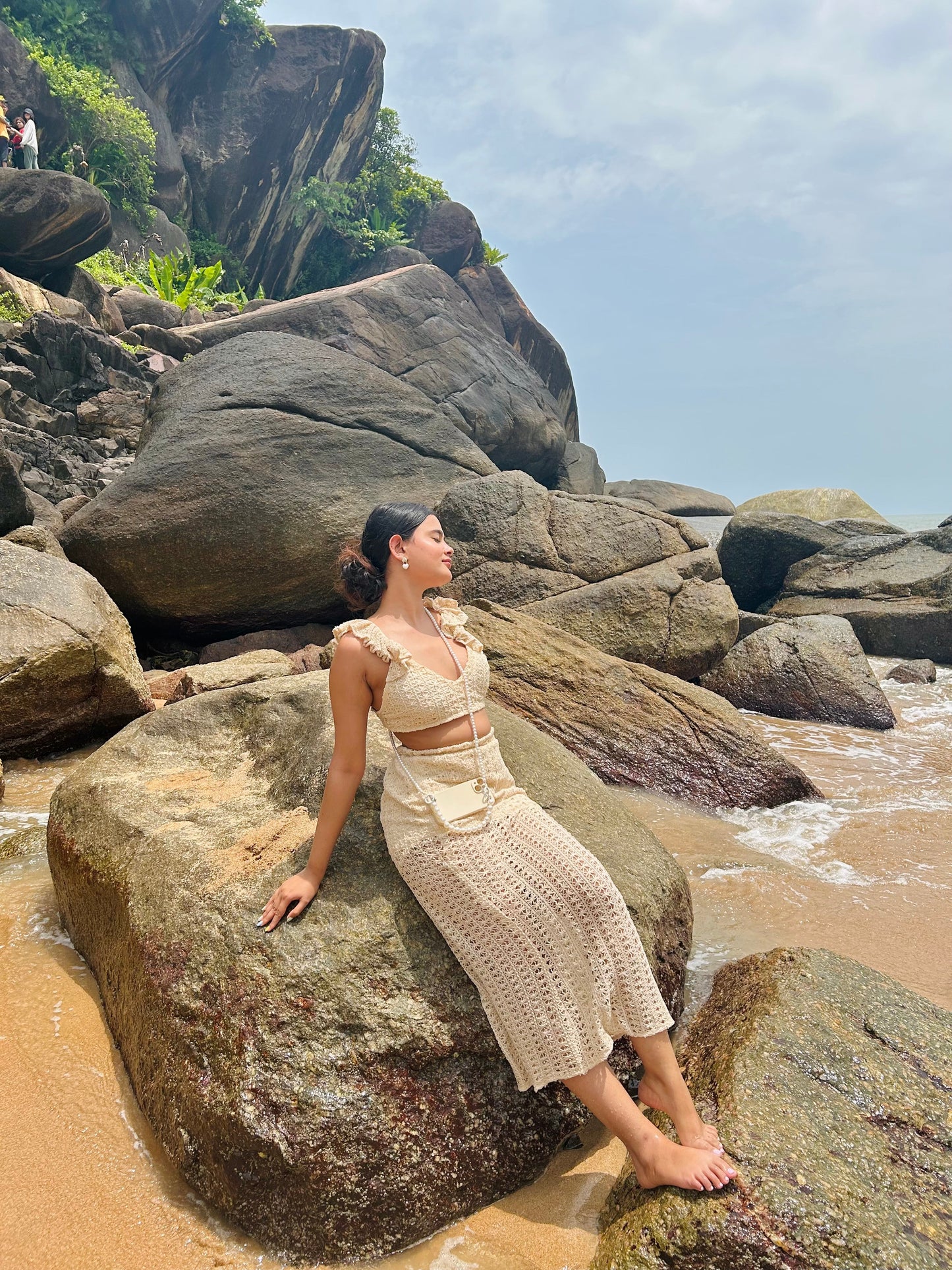 Stylera model in beige crochet two piece set sitting on coastal rocks and sand in a trendy seaside vacation outfit