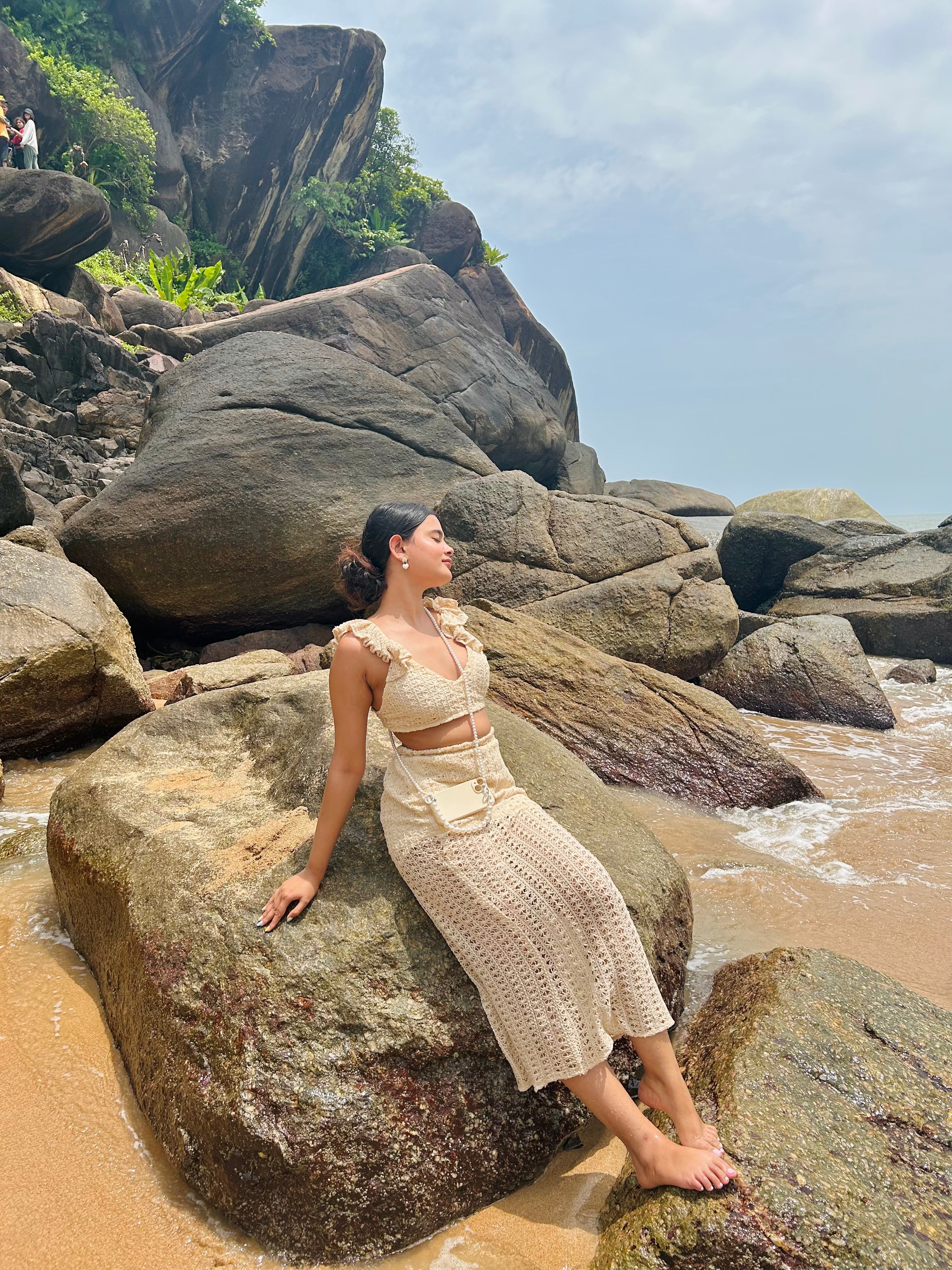Stylera model in beige crochet two piece set sitting on coastal rocks and sand in a trendy seaside vacation outfit