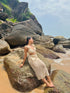 Stylera model in beige crochet two piece set sitting on coastal rocks and sand in a trendy seaside vacation outfit