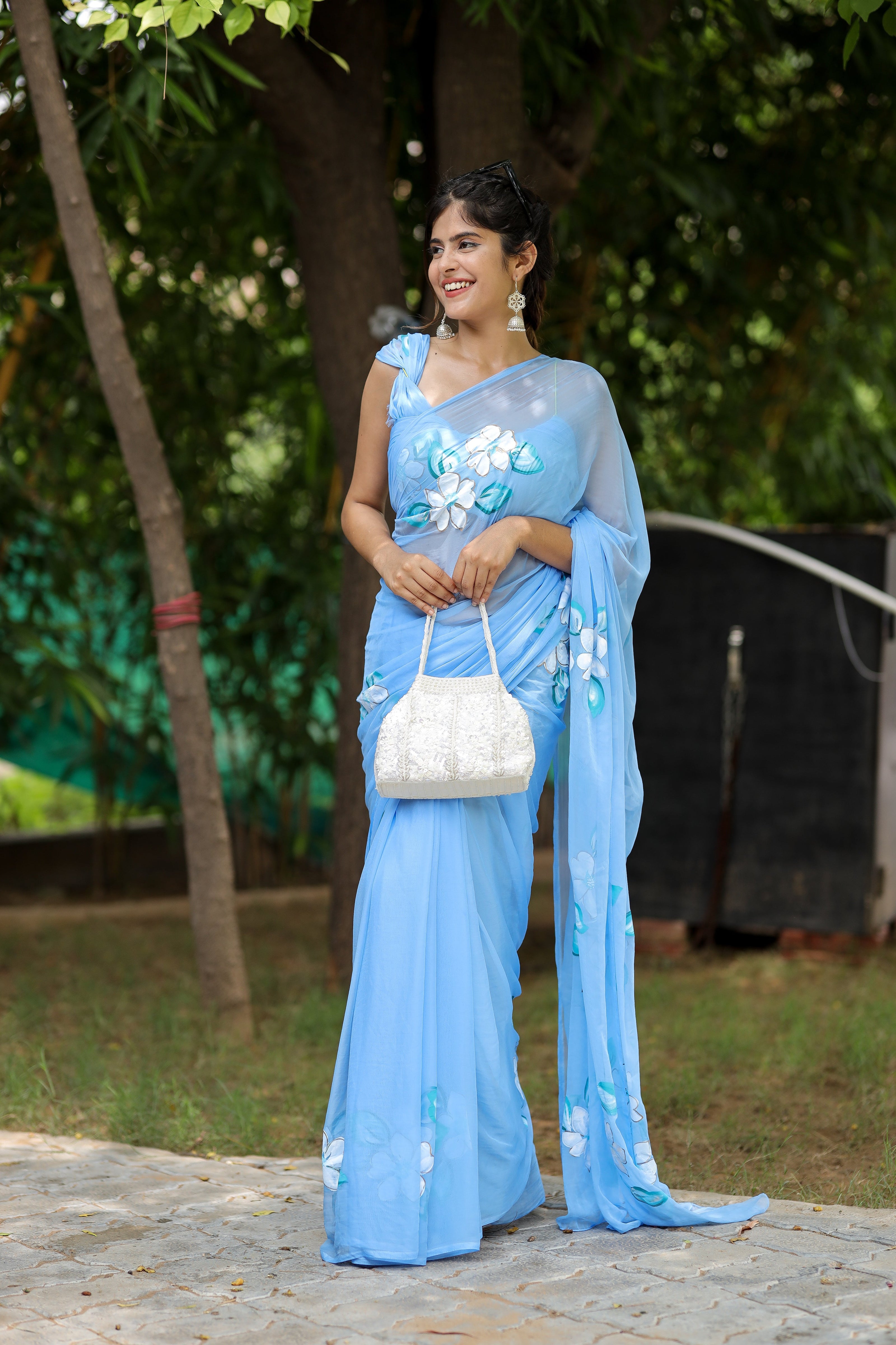 Elegant designer chiffon saree with floral painting in blue, styled with a white handbag and accessories