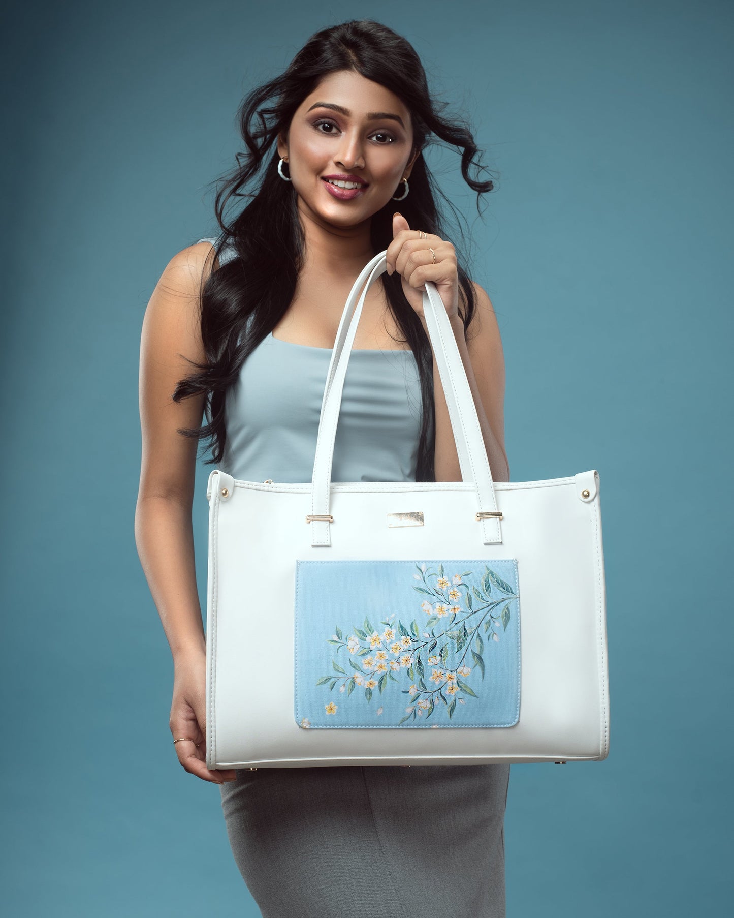 Model showcasing a large designer tote bag with floral painting on a blue background