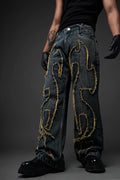 An image of a person wearing dark-wash, wide-leg denim jeans with a distinctive distressed design. The jeans feature large, flame-like patterns outlined with frayed, yellow-tinted fabric edges across the front of both legs. The person is also wearing a black tank top, black gloves, and chunky black boots, posing against a dark gray background.