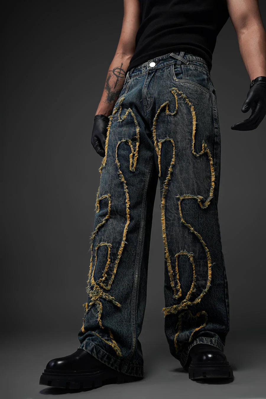 An image of a person wearing dark-wash, wide-leg denim jeans with a distinctive distressed design. The jeans feature large, flame-like patterns outlined with frayed, yellow-tinted fabric edges across the front of both legs. The person is also wearing a black tank top, black gloves, and chunky black boots, posing against a dark gray background.