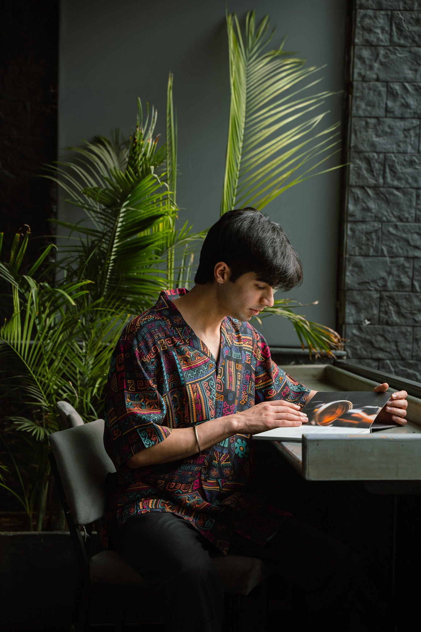 Earthy print oversized shirt for men featuring a vibrant design, worn while sitting at a table with plants in the background
