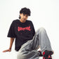 Model in Stylera black tee with red logo seated in baggy gray jeans and red black sneakers edgy graphic tee for men and women