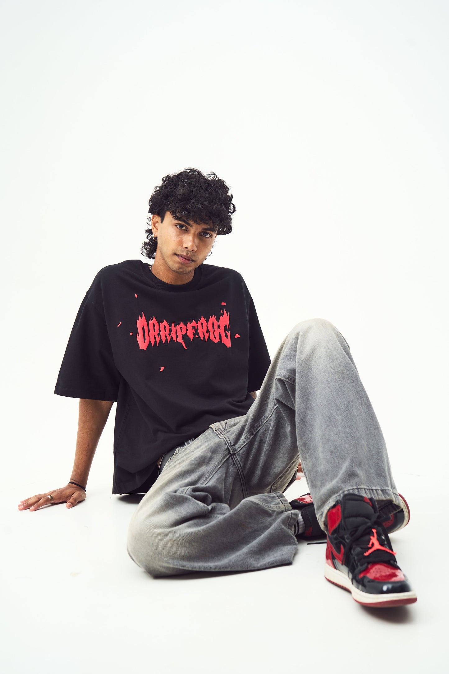 Model in Stylera black tee with red logo seated in baggy gray jeans and red black sneakers edgy graphic tee for men and women