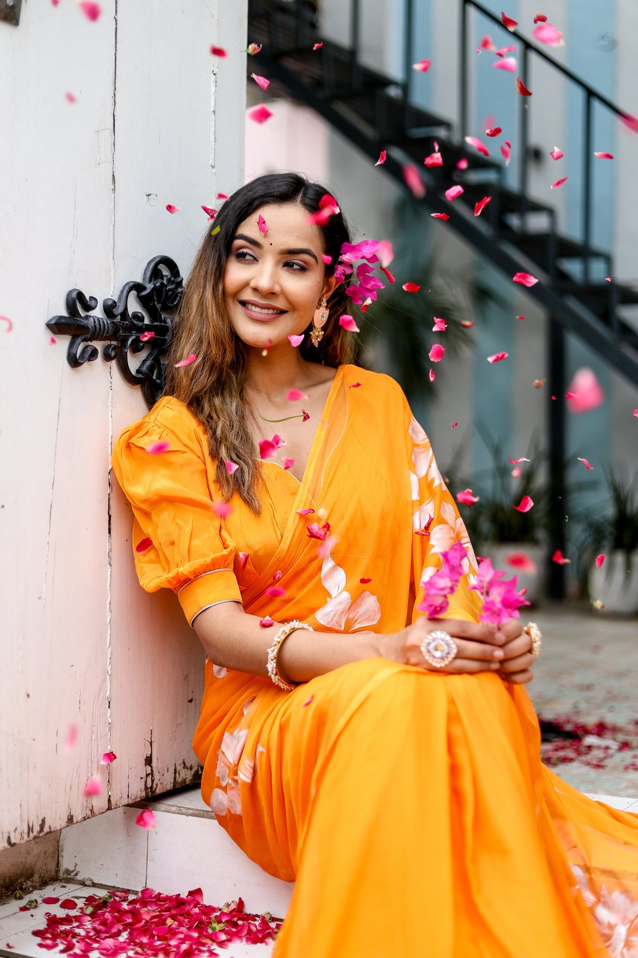Elegant woman sitting amid rose petals in a handpainted chiffon saree with intricate floral designs