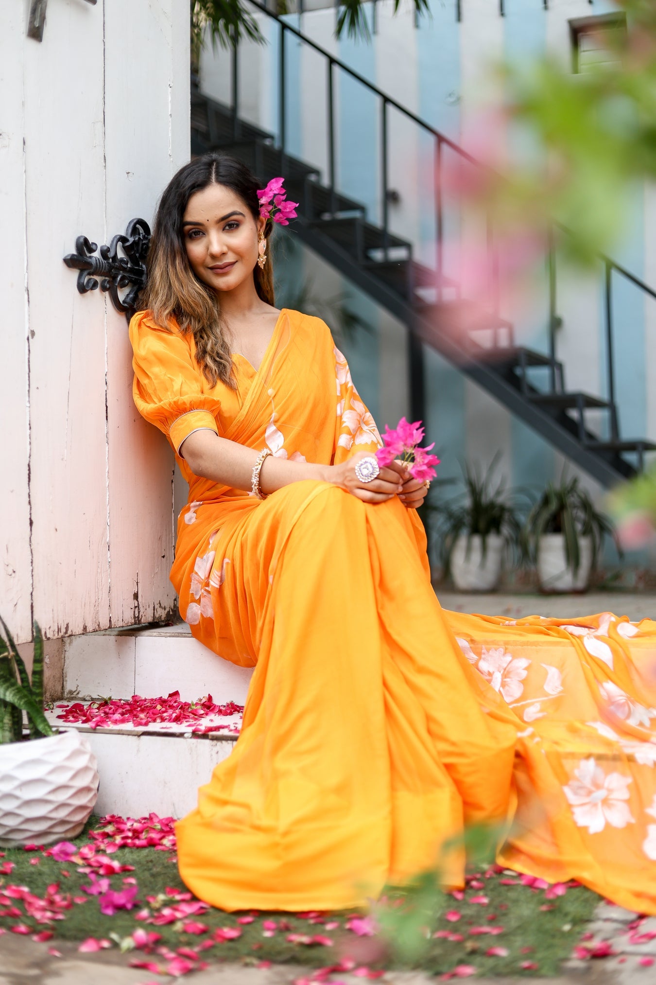 Elegant woman wearing a handpainted chiffon saree in vibrant orange with floral designs, seated in a garden