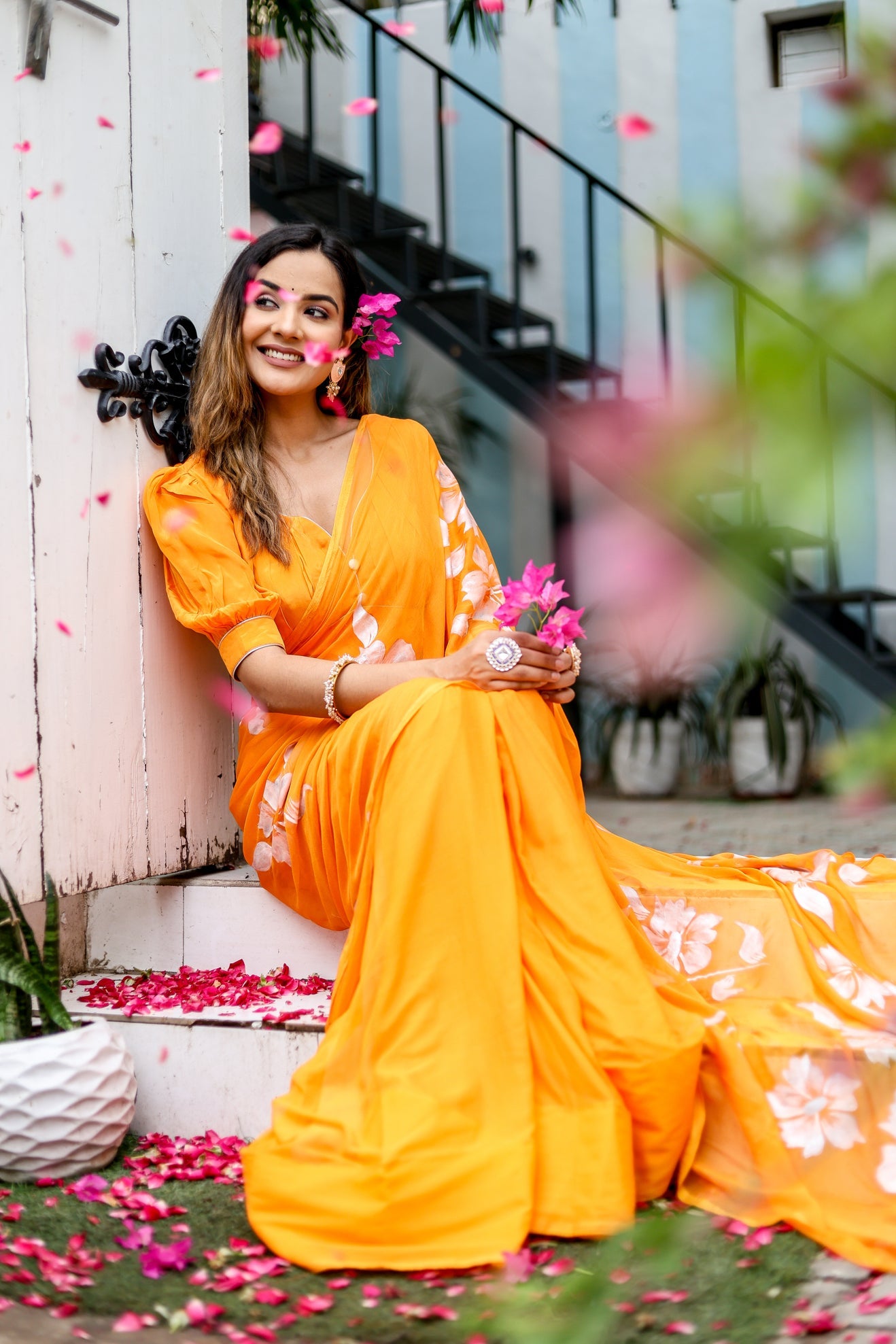 Elegant model wearing a vibrant handpainted chiffon saree in orange with floral designs, seated outdoors