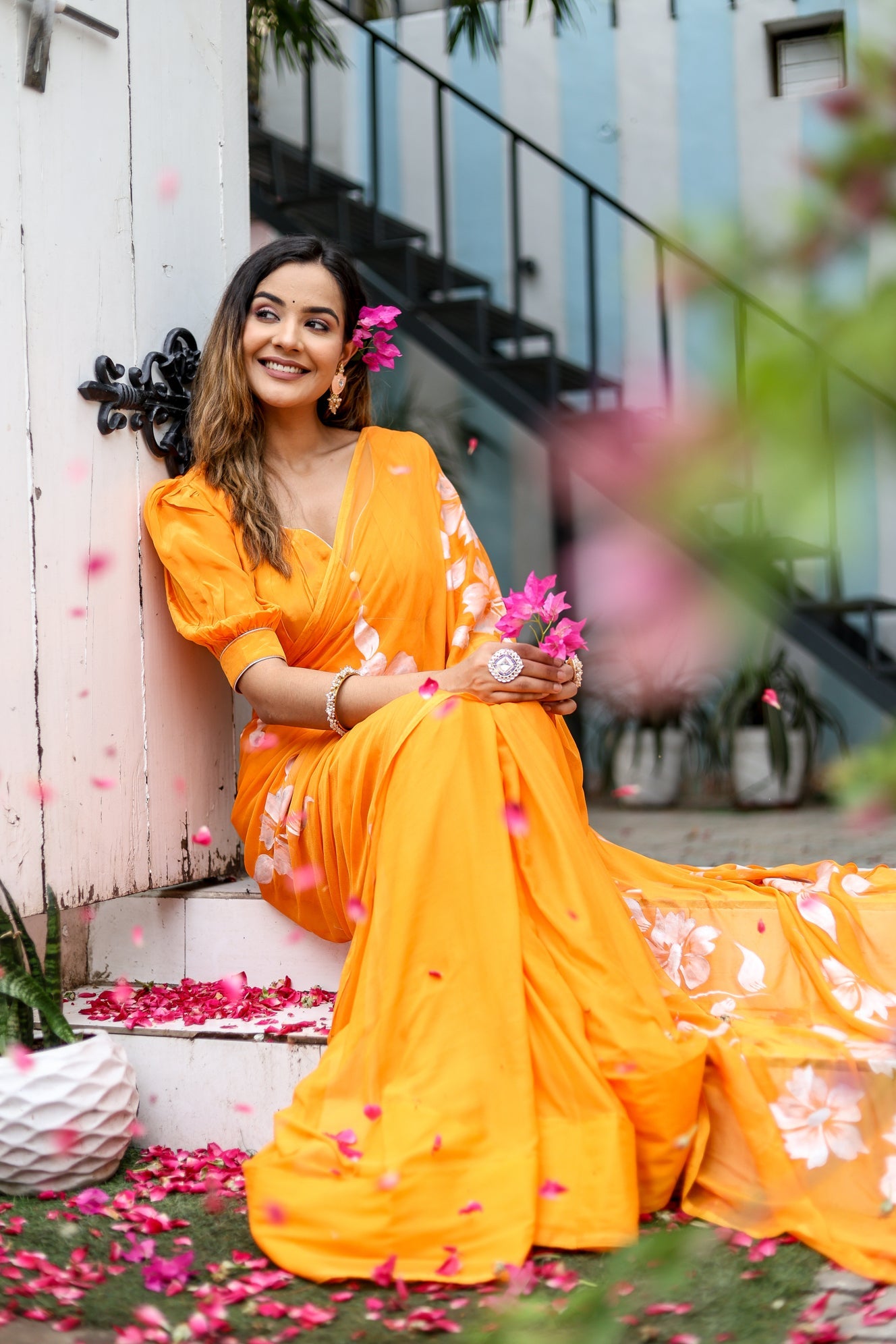 Bright orange handpainted chiffon saree with floral designs, worn elegantly by a smiling woman