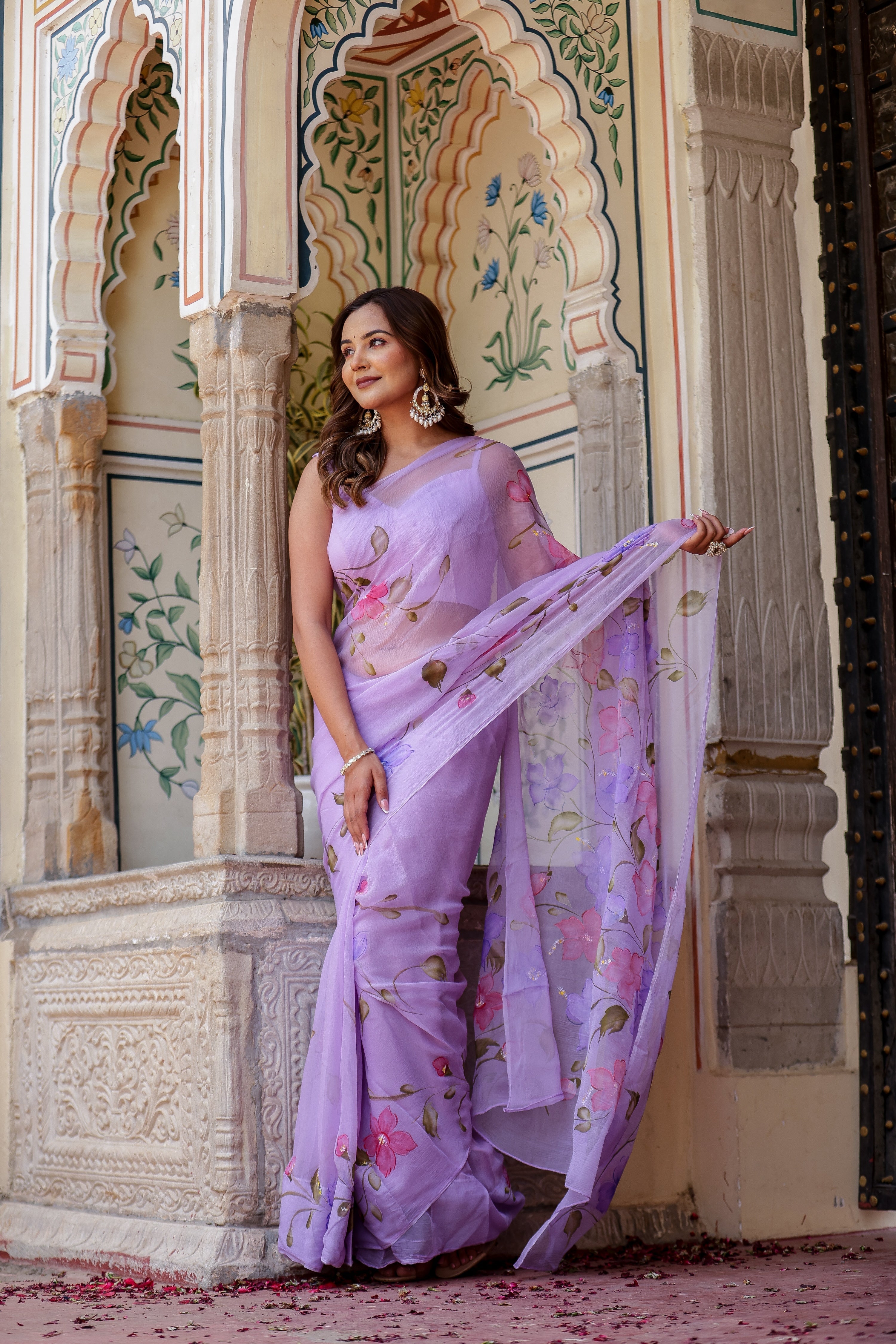 Elegant woman in a handpainted lavender chiffon saree with floral patterns, set against a decorative backdrop