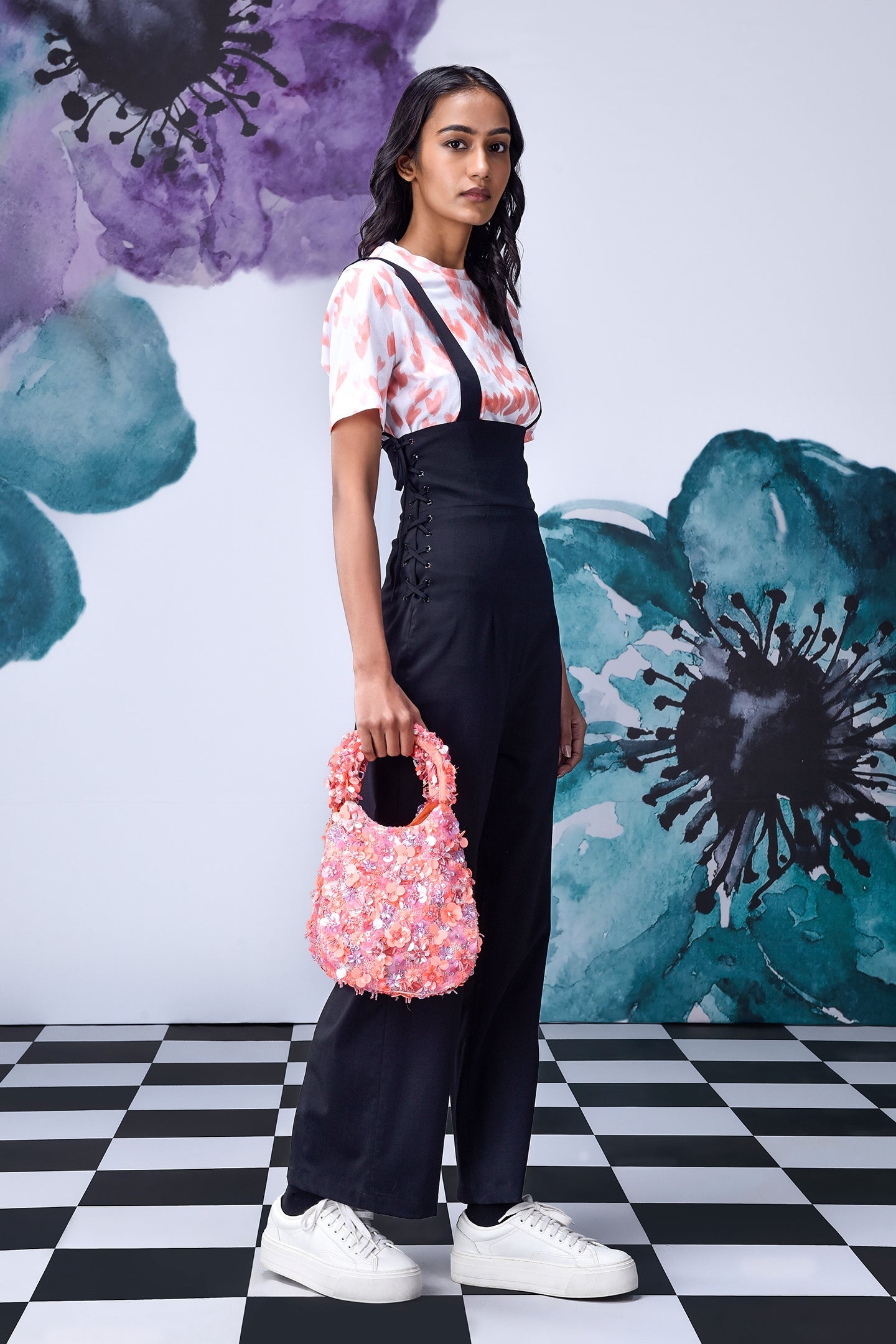 Stylera model wearing black dungaree styled high waist lace up overalls with pink floral handbag and white sneakers