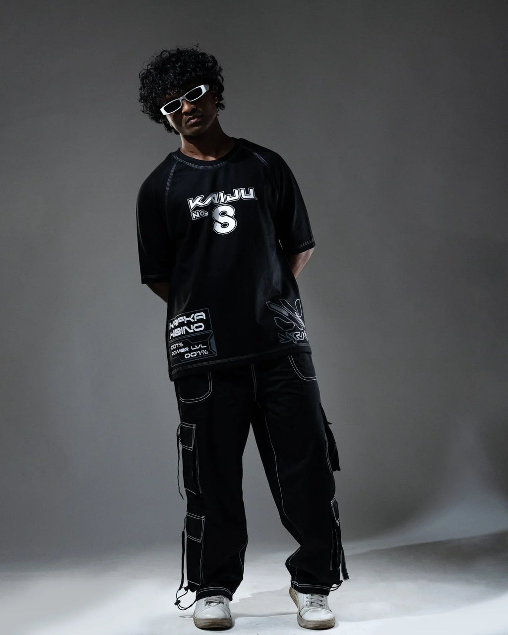Person wearing a black t-shirt with white text and graphics, dark cargo pants, and white sneakers on a gray background.