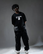 Person wearing a black t-shirt with white text and graphics, dark cargo pants, and white sneakers on a gray background.
