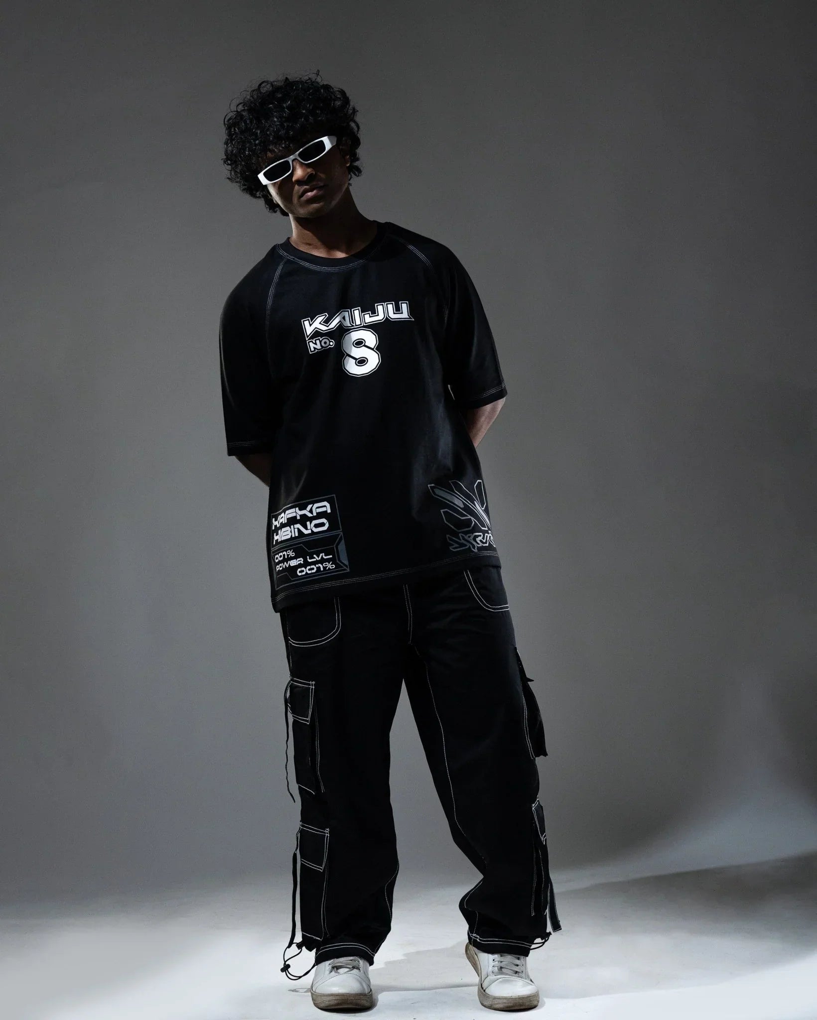 Person wearing a black t-shirt with white text and graphics, dark cargo pants, and white sneakers on a gray background.