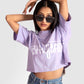 Young woman wearing a lavender oversized crop top with white text and sunglasses posing with arms raised