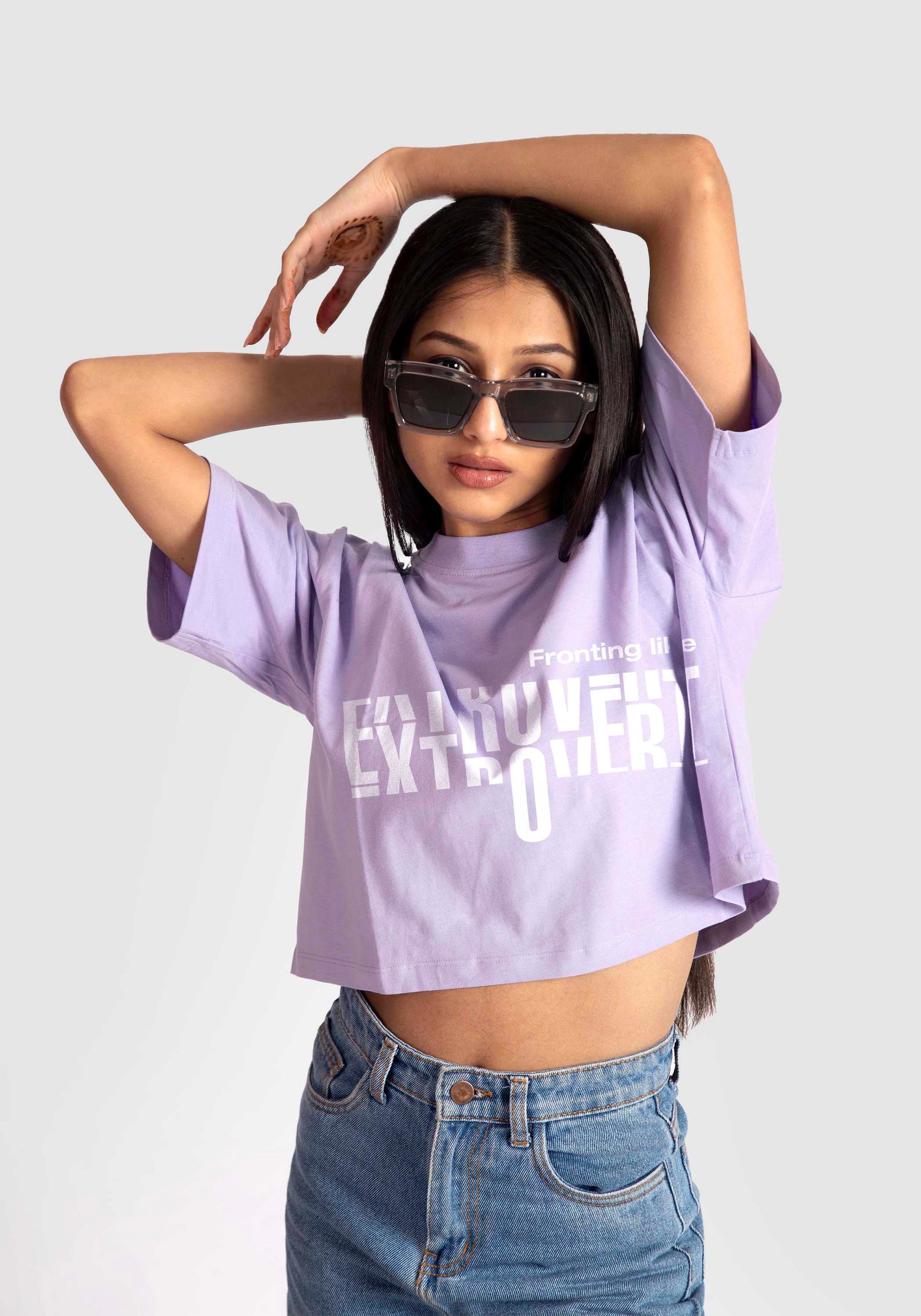 Young woman wearing a lavender oversized crop top with white text and sunglasses posing with arms raised
