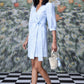 Model wearing a light blue wrap dress with three quarter sleeves and waist tie holding woven bag on checkered floor