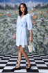 Model wearing a light blue wrap dress with three quarter sleeves and waist tie holding woven bag on checkered floor
