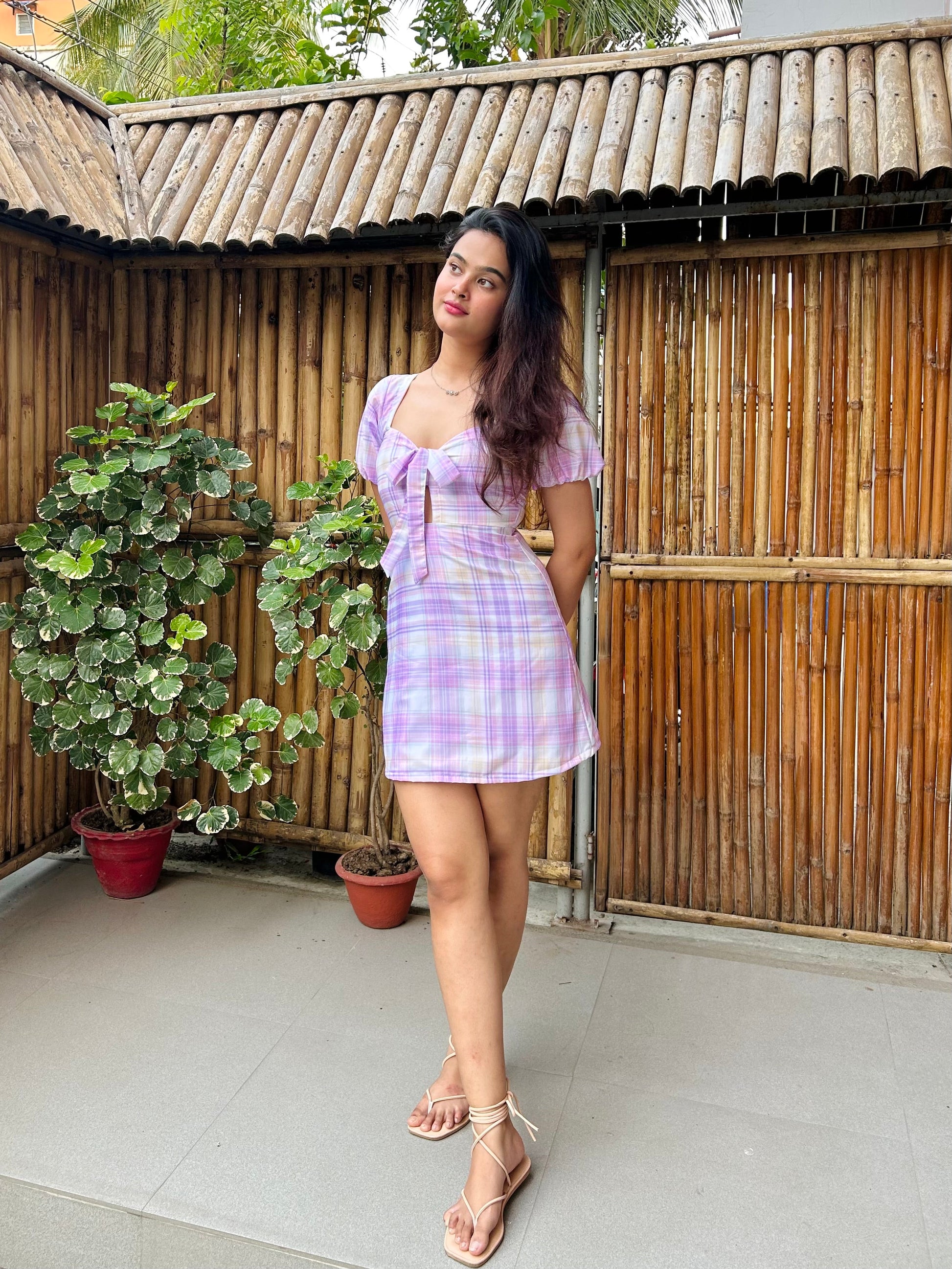 Model wearing Stylera trendy short sleeve mini dress in lilac plaid with tie front and sandals on bamboo patio