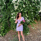 Model wearing trendy short sleeve mini dress purple gingham holding pink roses by white flowering hedge