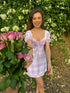Young woman smiling in Stylera trendy short sleeve mini dress pink plaid tie front puff sleeves holding roses outdoors
