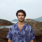Model wearing a men's abstract blue oversize shirt standing on rocky shoreline with a scenic background