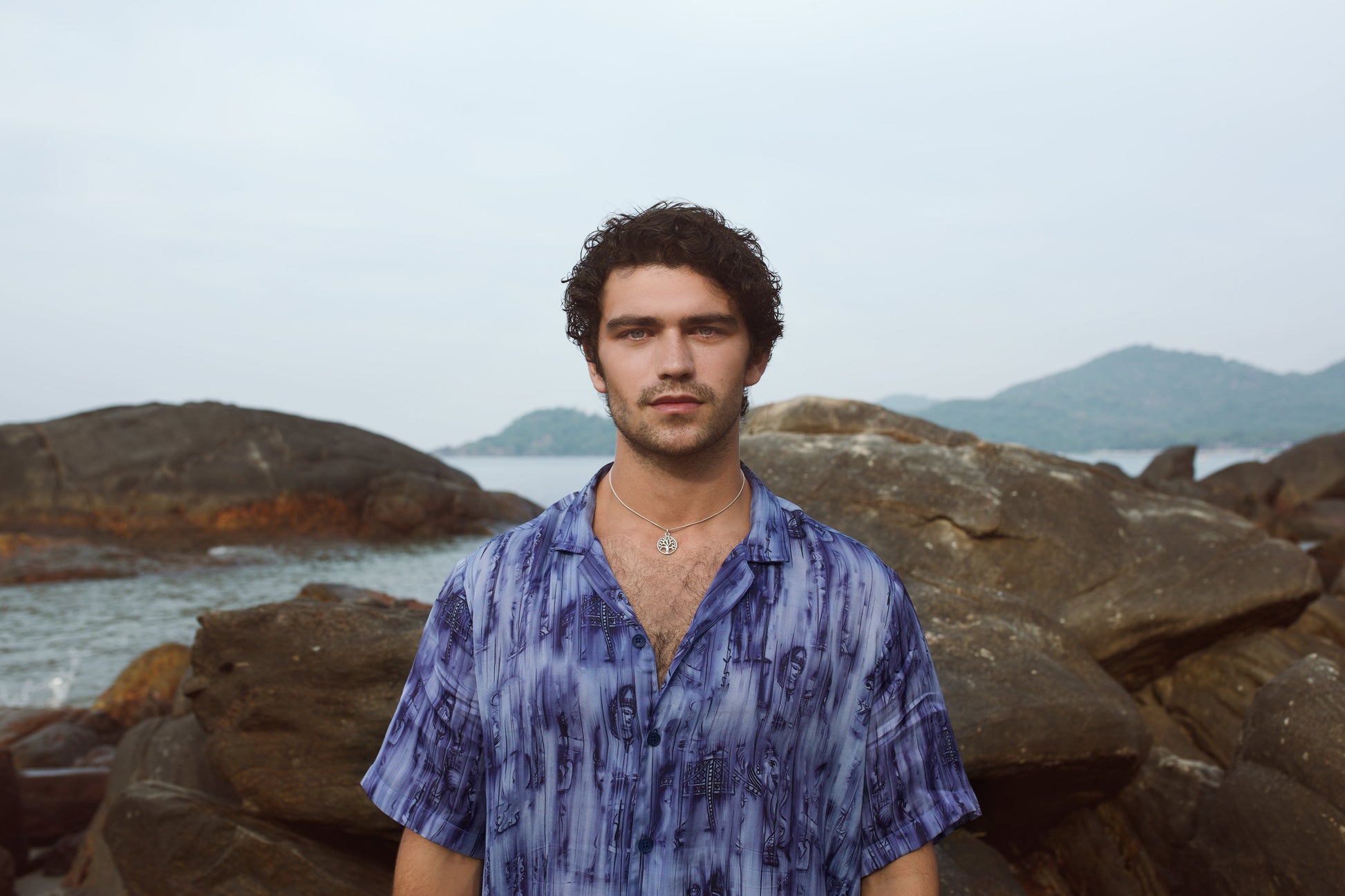 Model wearing a men's abstract blue oversize shirt standing on rocky shoreline with a scenic background
