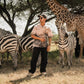 Man wearing men's printed casual button up shirt and sunglasses standing among zebras and giraffe in African savanna