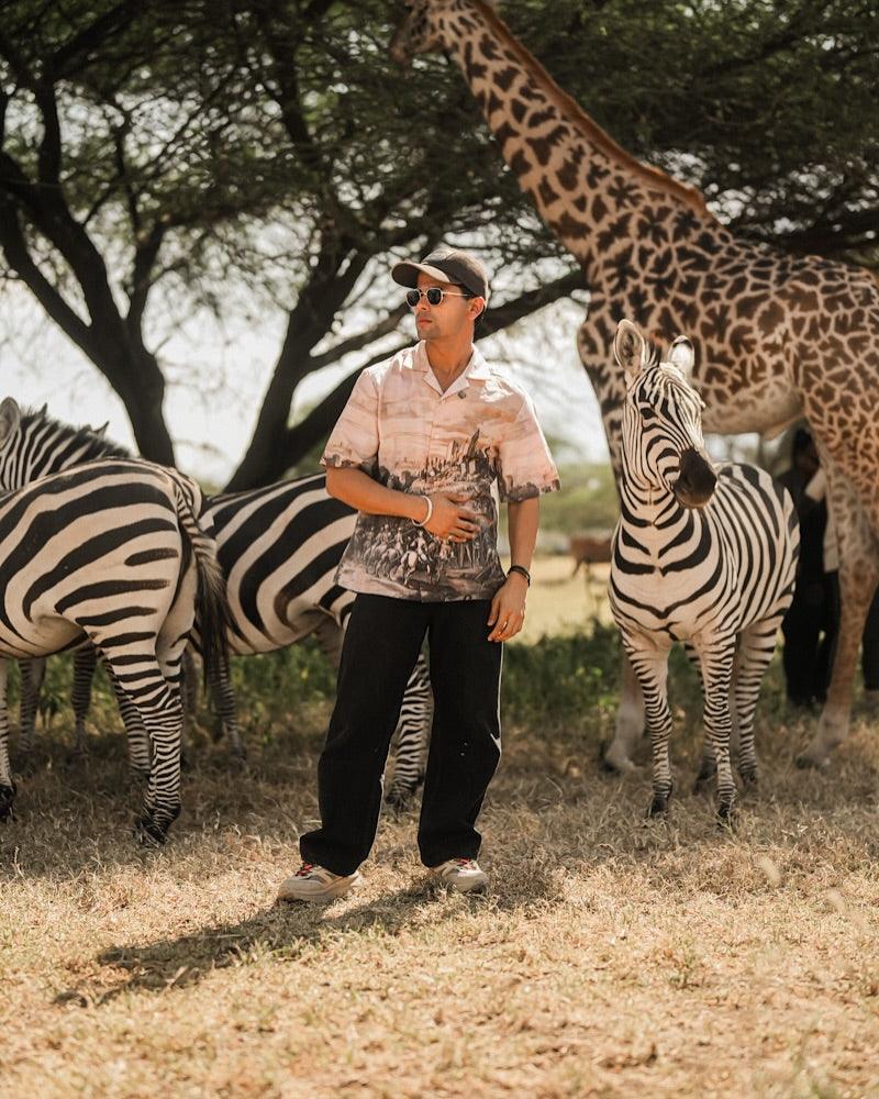 Man wearing men's printed casual button up shirt and sunglasses standing among zebras and giraffe in African savanna