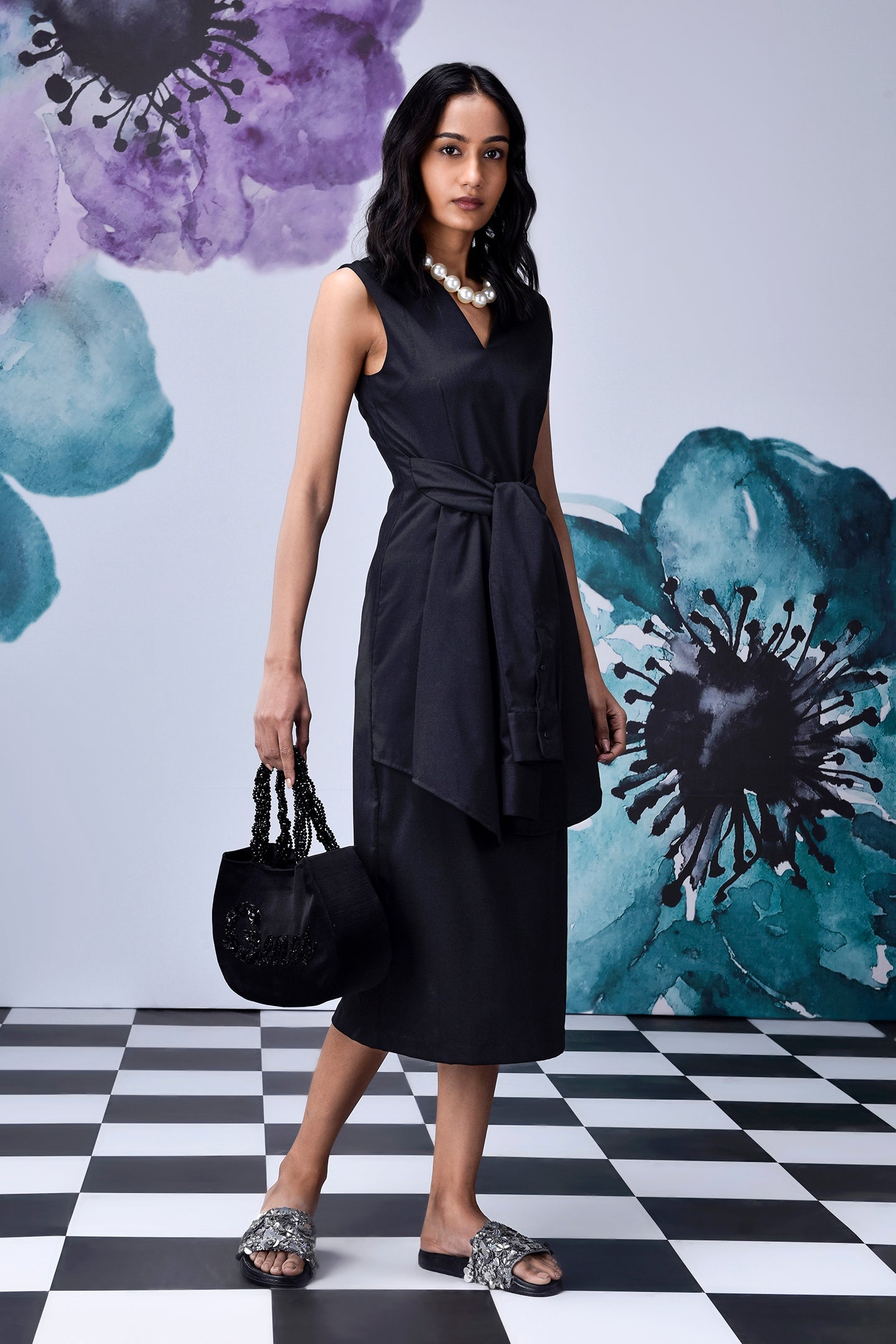 Stylera model wearing midi black dress with belt side tie V neck sleeveless with pearl necklace and black handbag