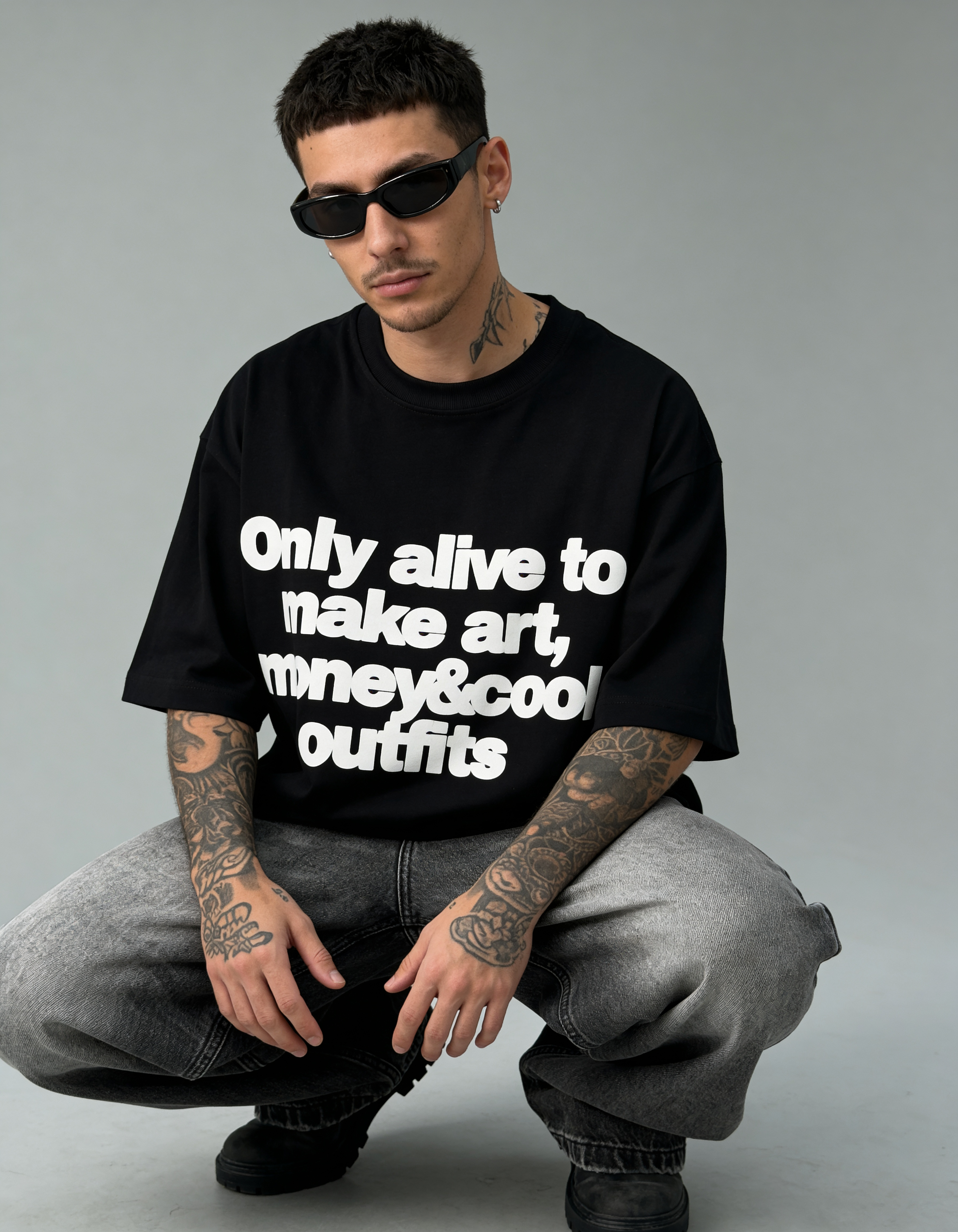 Man wearing a black t-shirt with text, sitting on the ground against a gray background